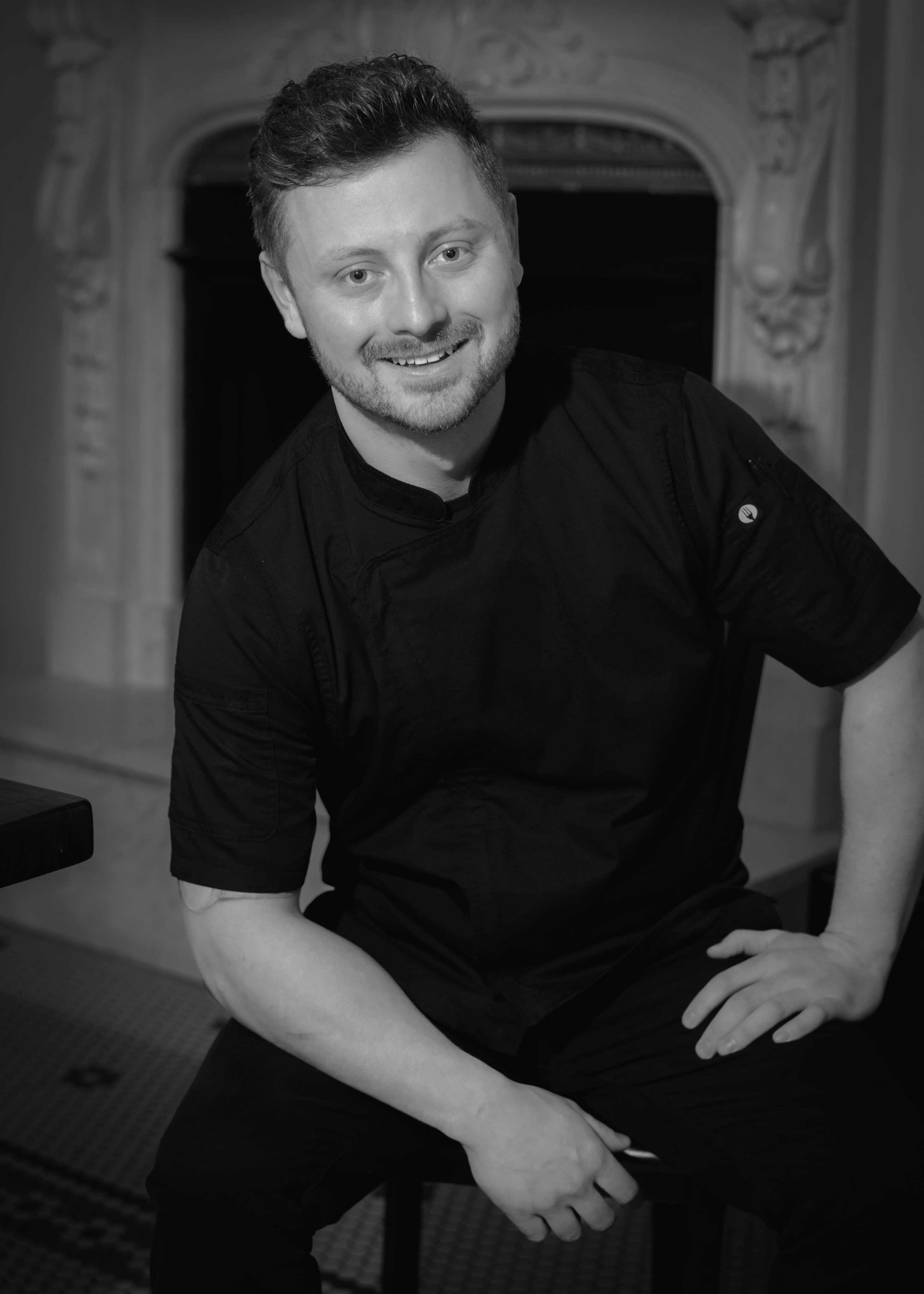 Smiling man with short hair and beard wearing a black chef coat, sitting indoors with one hand on his knee.