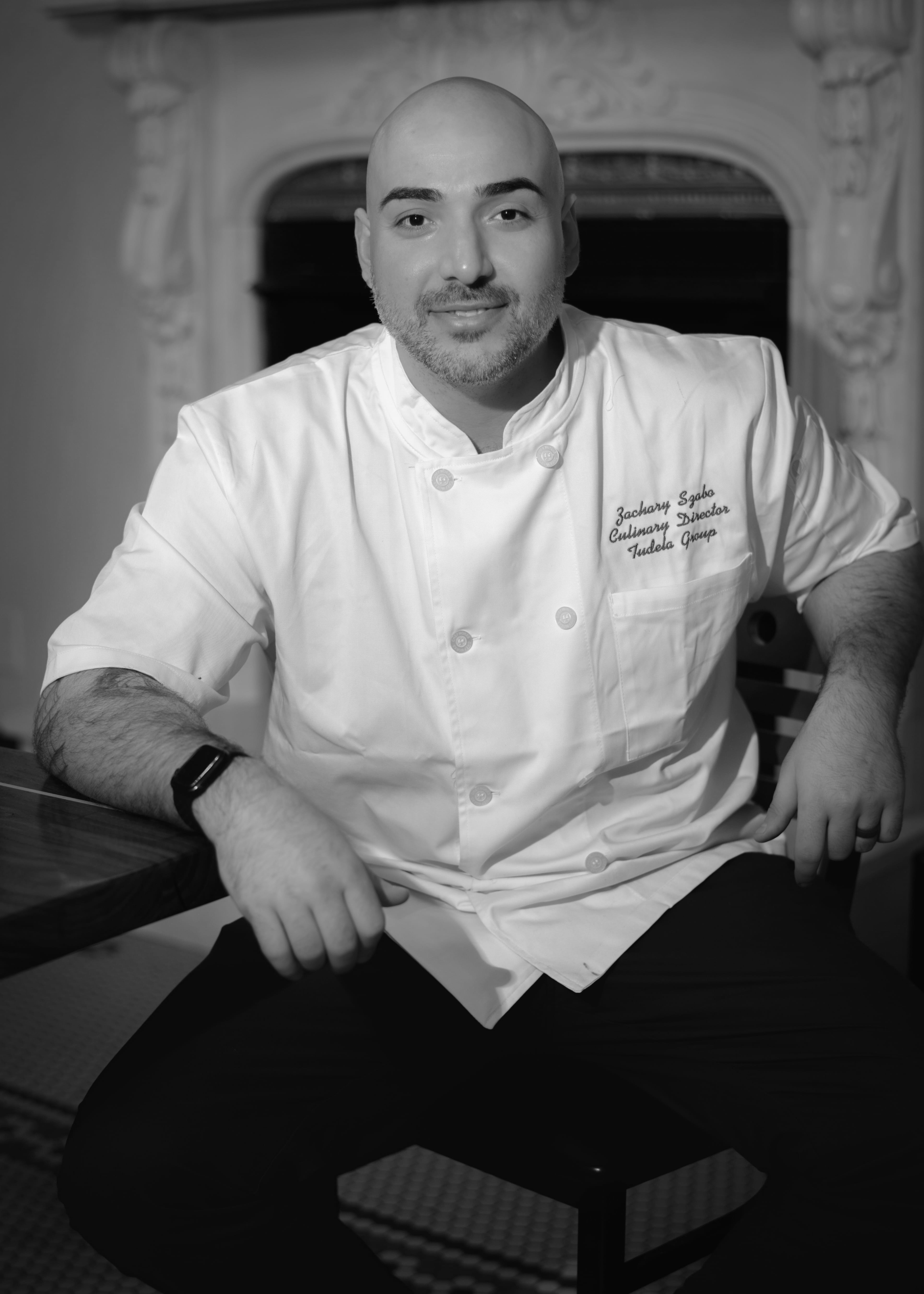 Portrait of a bald man with a beard wearing a chef's jacket embroidered with 'Zachary Szabo Culinary Director Tudela Group,' sitting at a table.