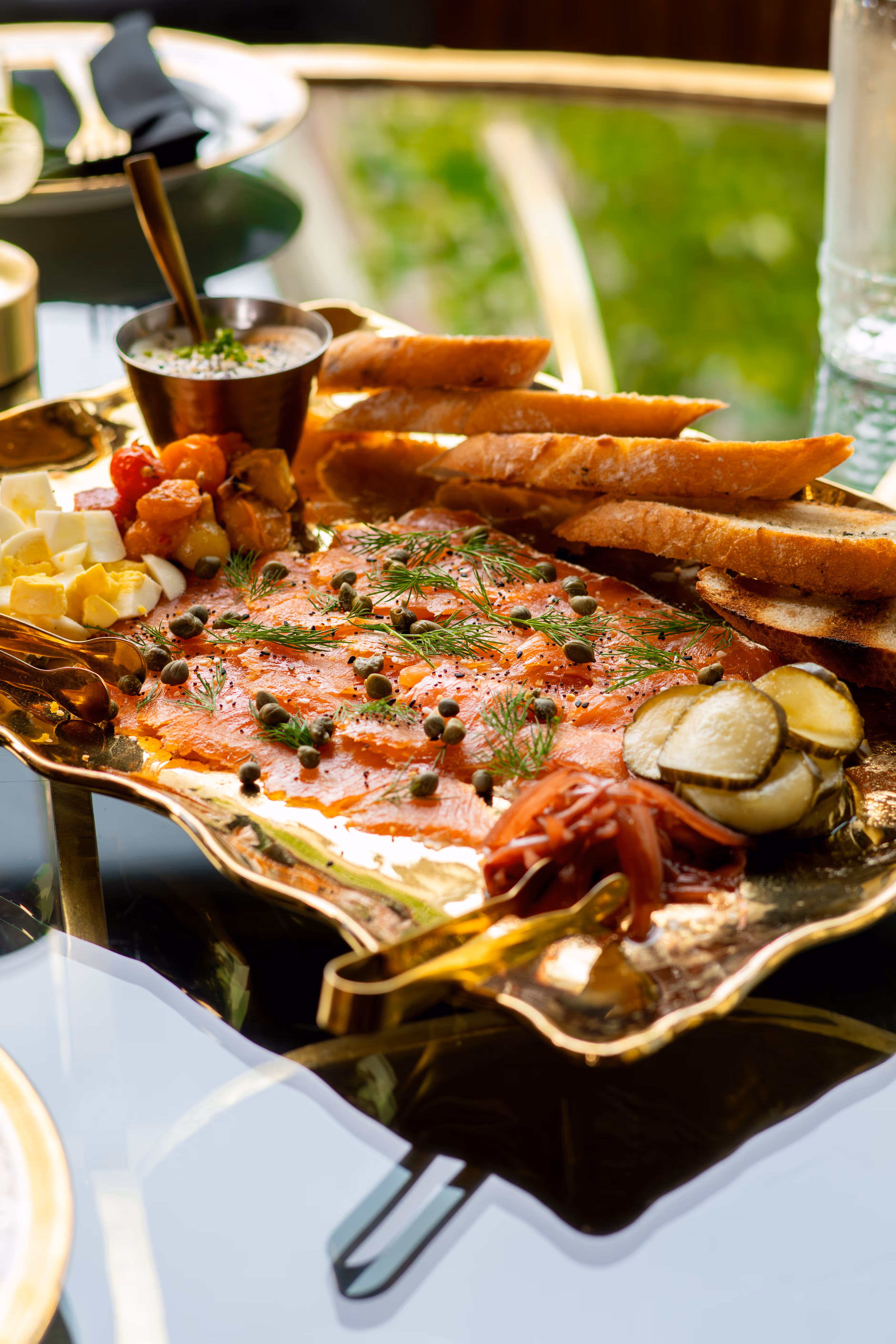 Platter with smoked salmon garnished with capers and dill, baguette slices, pickles, diced boiled eggs, and sauce.