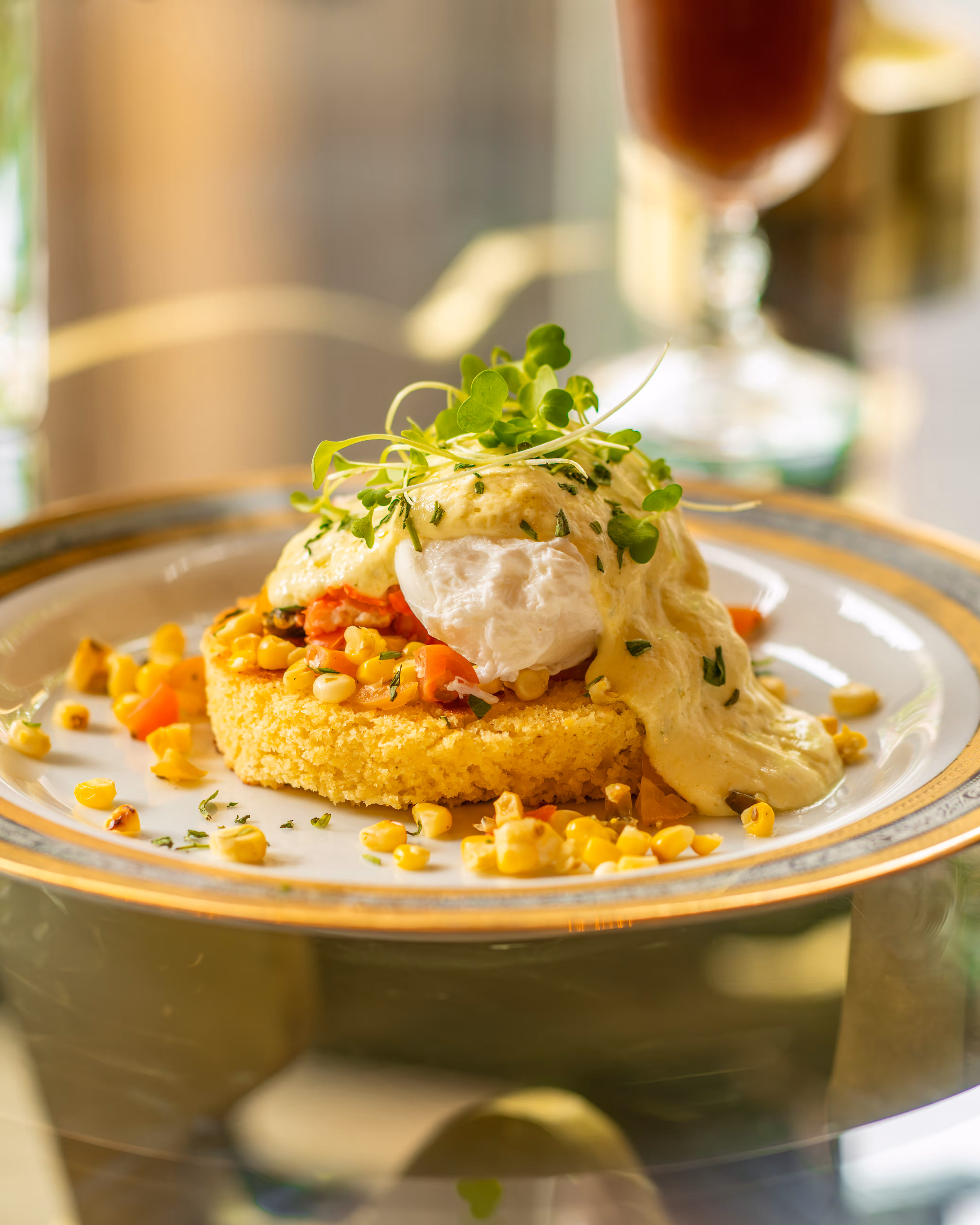 Poached egg on a round corn cake topped with creamy sauce, microgreens, and corn kernels on a decorative plate.