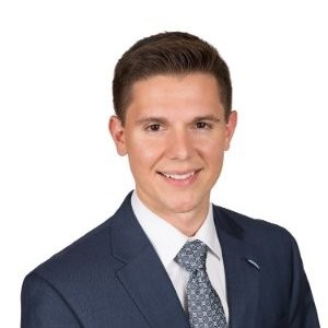 Young man wearing a navy blue suit, white shirt, and patterned tie, smiling against a white background.