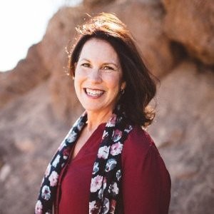 Smiling woman with dark hair wearing a maroon top and floral scarf standing outdoors with rocky background.