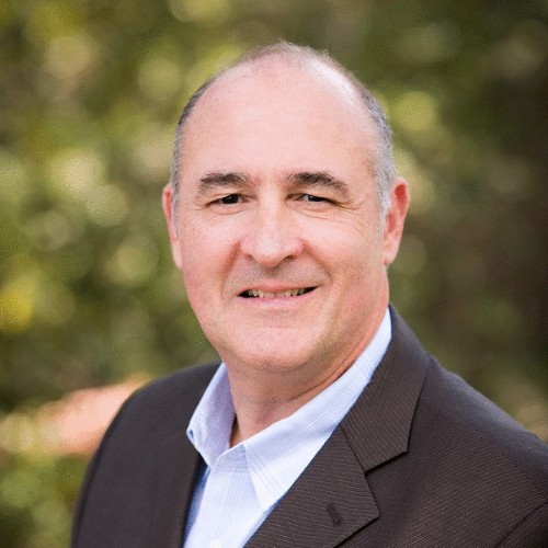 Smiling middle-aged man in a brown blazer and light blue shirt with a blurred outdoor background.