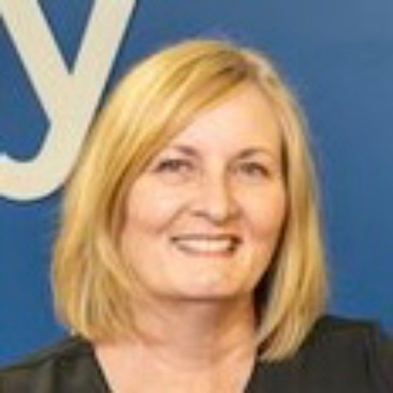 Smiling woman with shoulder-length blonde hair wearing a black top against a blue background.