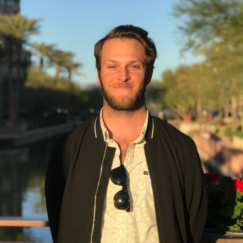 Smiling man with short brown hair and beard wearing a light patterned shirt and black jacket, standing outdoors with trees and water in the background.