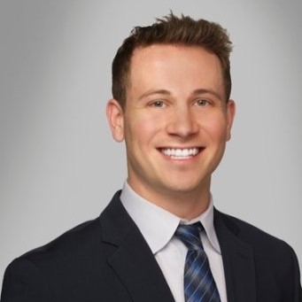 Smiling man with short brown hair wearing a dark suit, white shirt, and blue striped tie against a gray background.