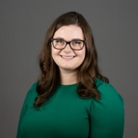 Smiling woman with long brown hair and glasses wearing a green top against a gray background.