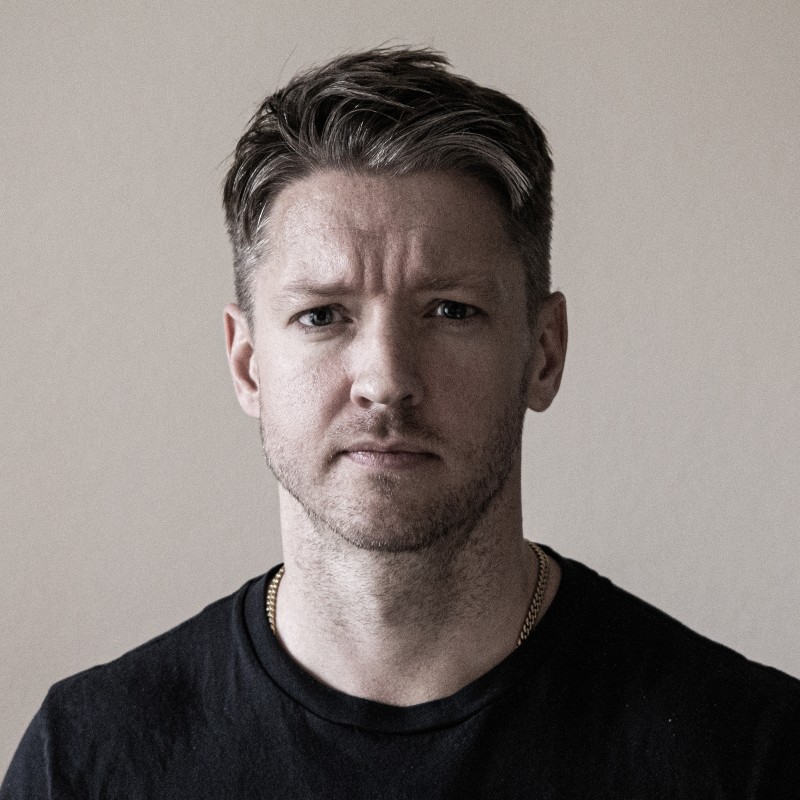 Serious man with short hair and light beard wearing a black shirt and gold chain against a plain background.