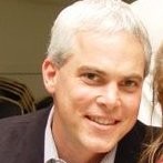 Smiling middle-aged man with short gray hair wearing a dark jacket and light-colored shirt.
