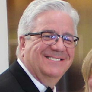 Smiling elderly man with white hair and glasses wearing a dark suit.