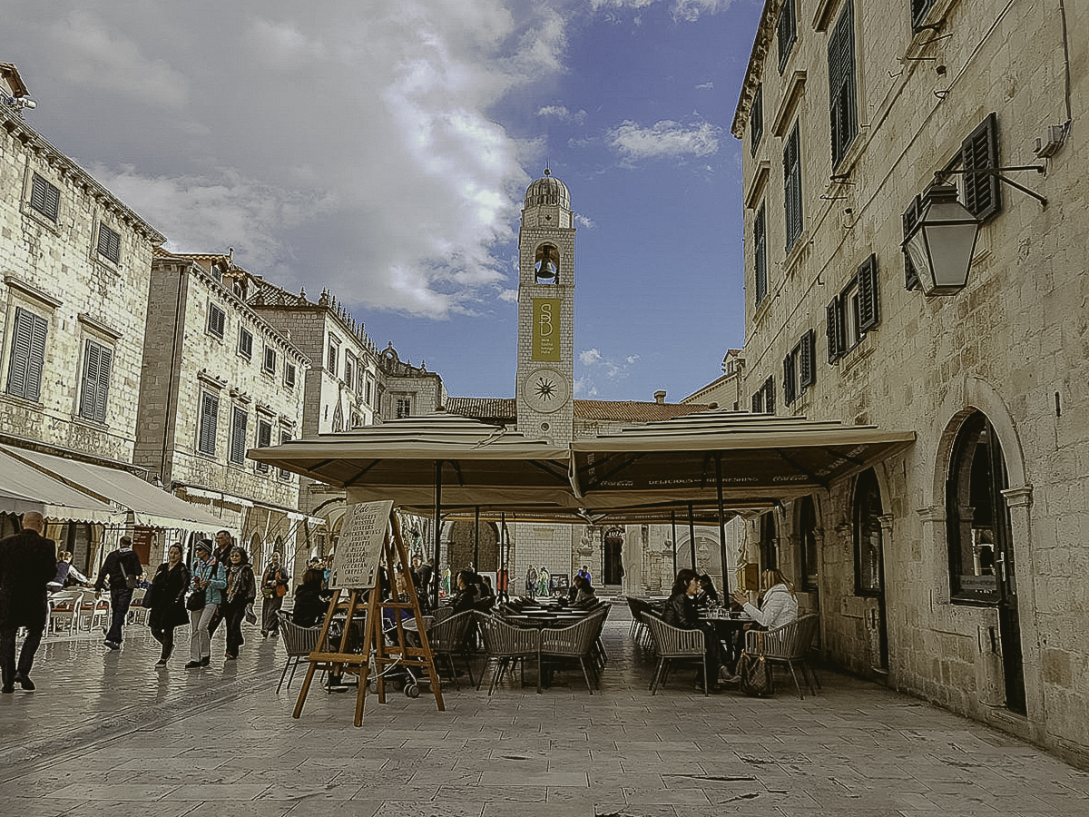 People sit and walk around an outdoor café in a historic European square.