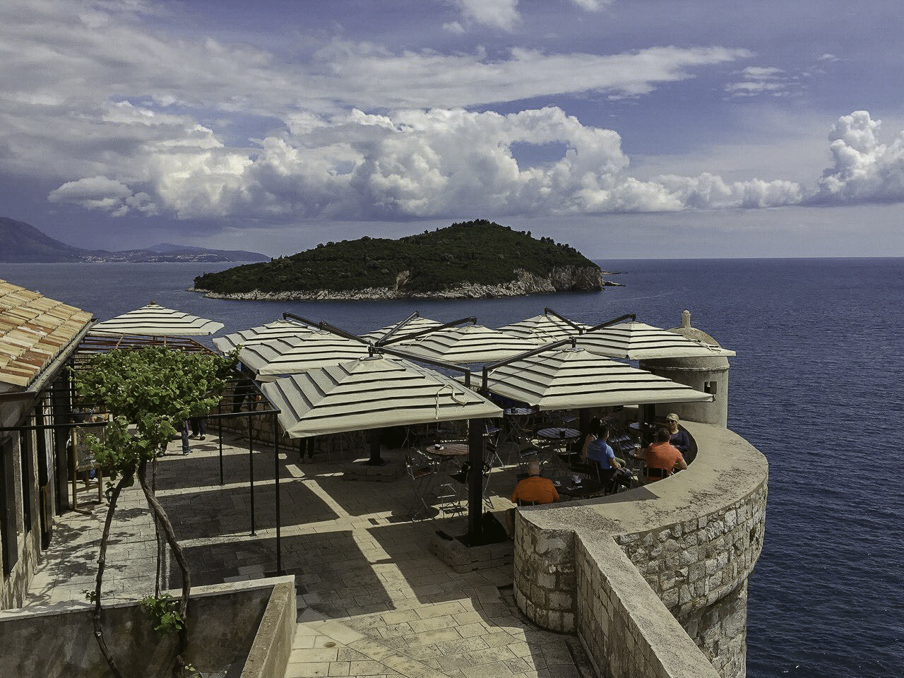 Outdoor café with visitors under striped umbrellas, overlooking the sea and a distant island.