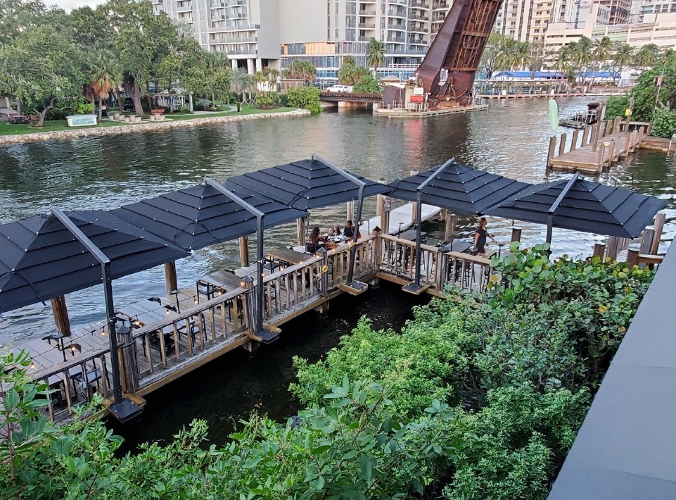A row of empty outdoor tables and chairs under black awnings by a river.