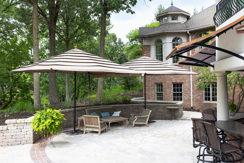 Patio with striped umbrellas, cushioned wooden chairs, and a stone wall near a large house.