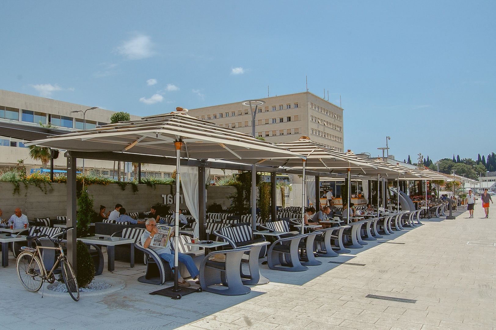 People sit at outdoor café tables under umbrellas, reading newspapers and chatting on a sunny day.