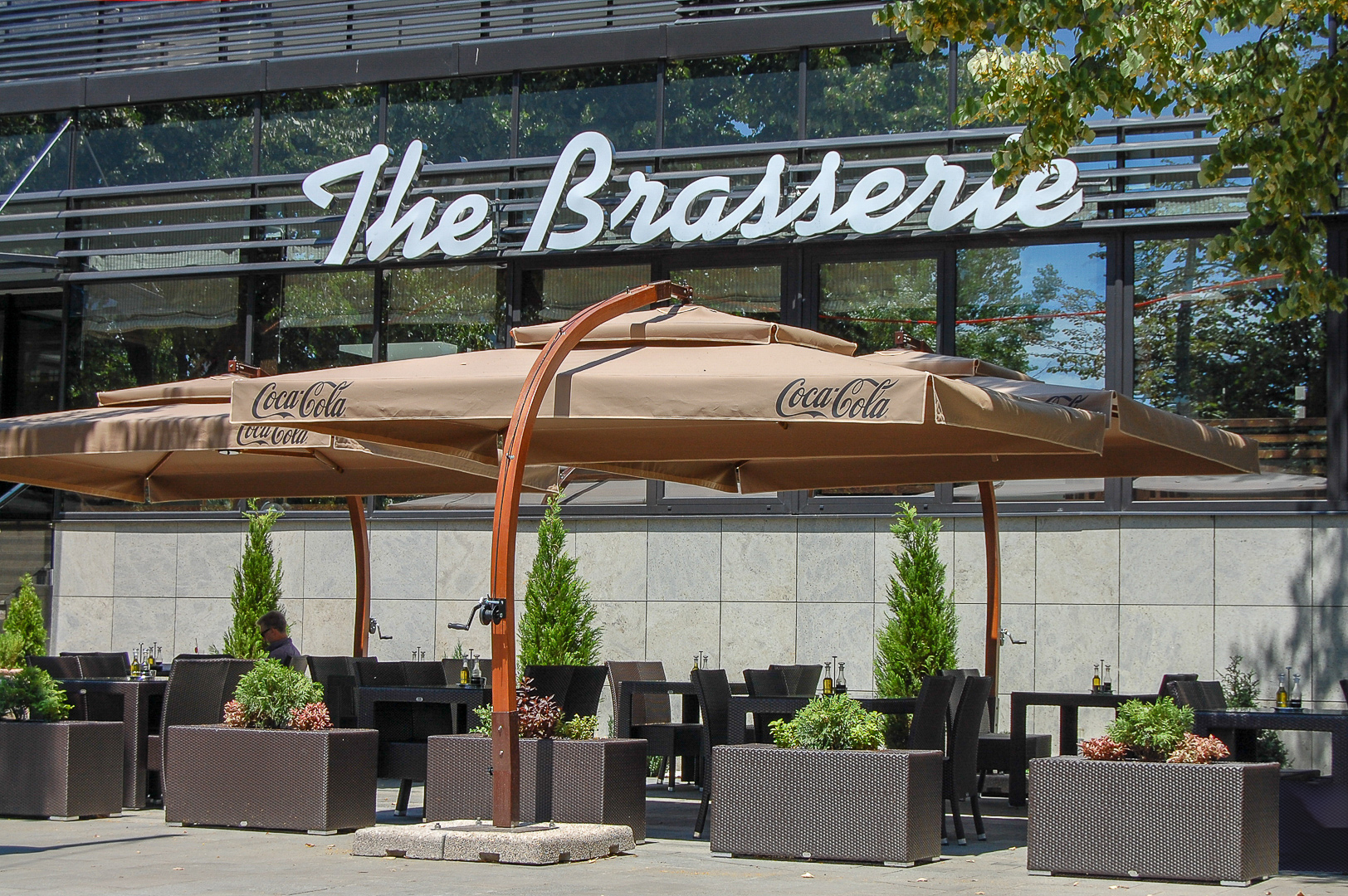 Outdoor restaurant seating with brown umbrellas and greenery in front of a building with "The Brasserie" sign.