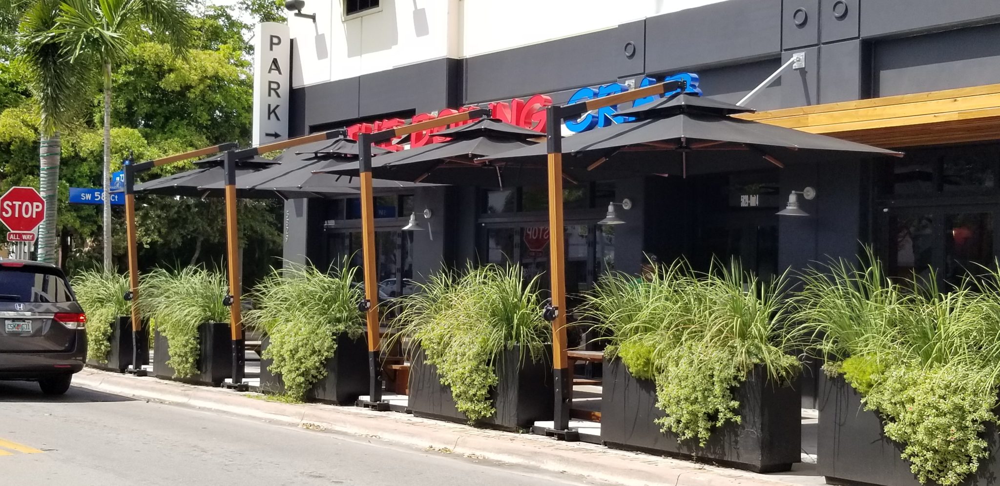 Restaurant patio with tables and chairs under black umbrellas, surrounded by tall potted plants.