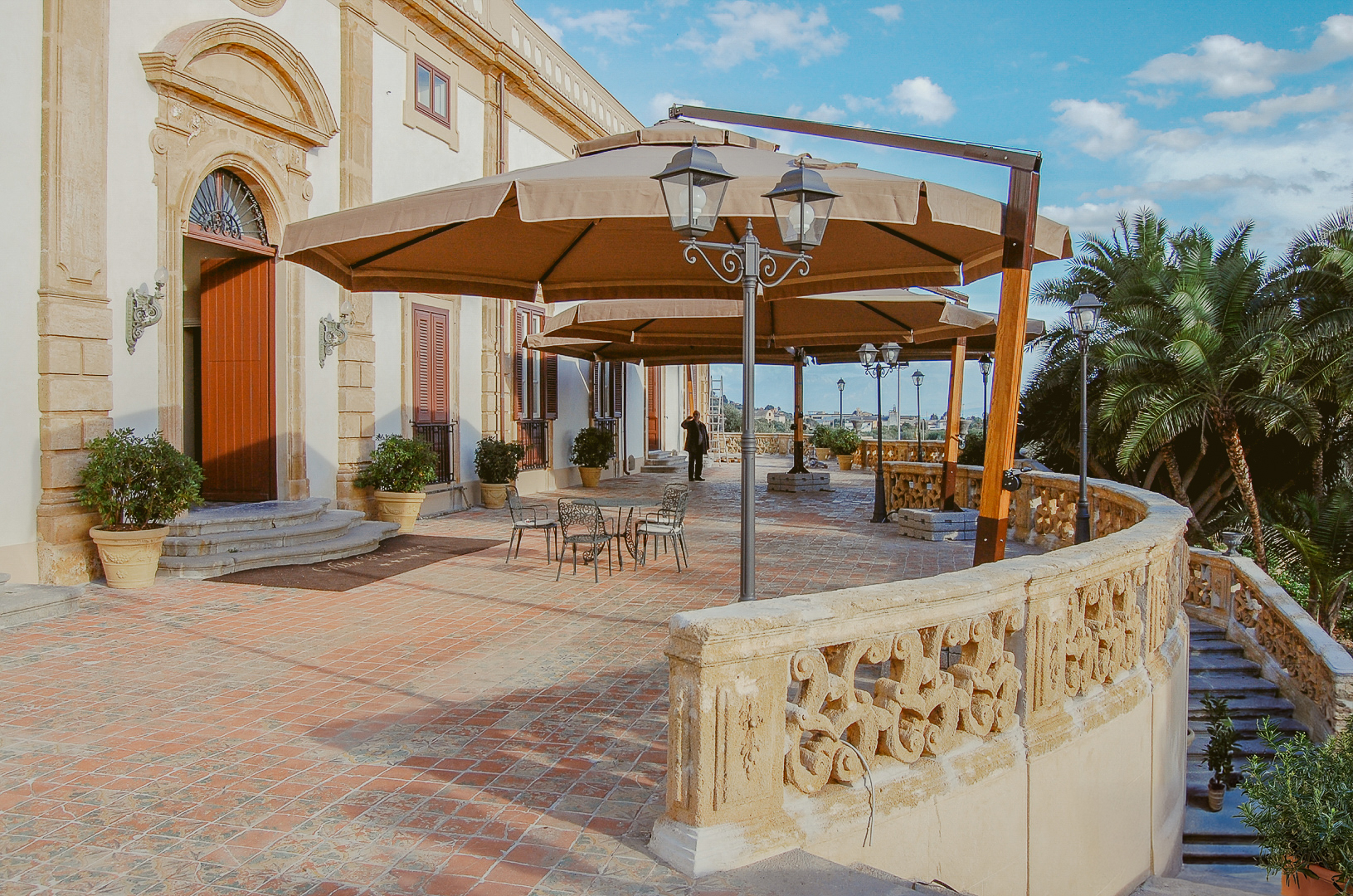 A person walks on a patio with tables, chairs, and large umbrellas near a building.