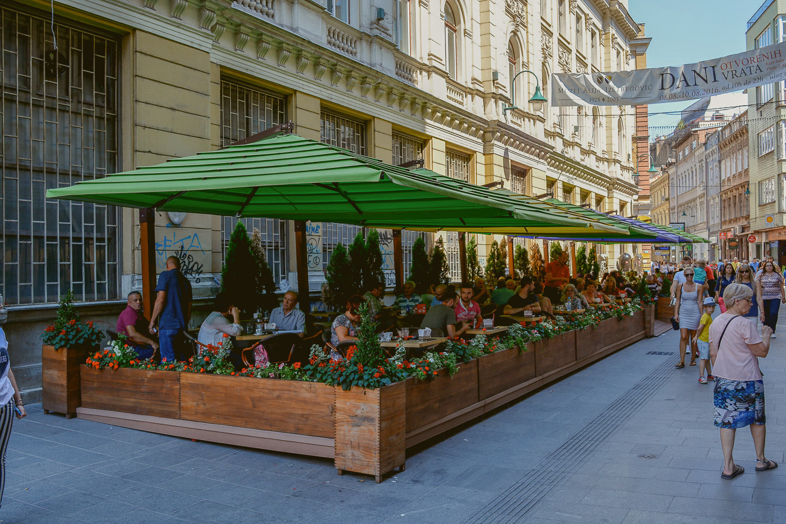 People are sitting and eating at outdoor tables under green umbrellas on a busy street.