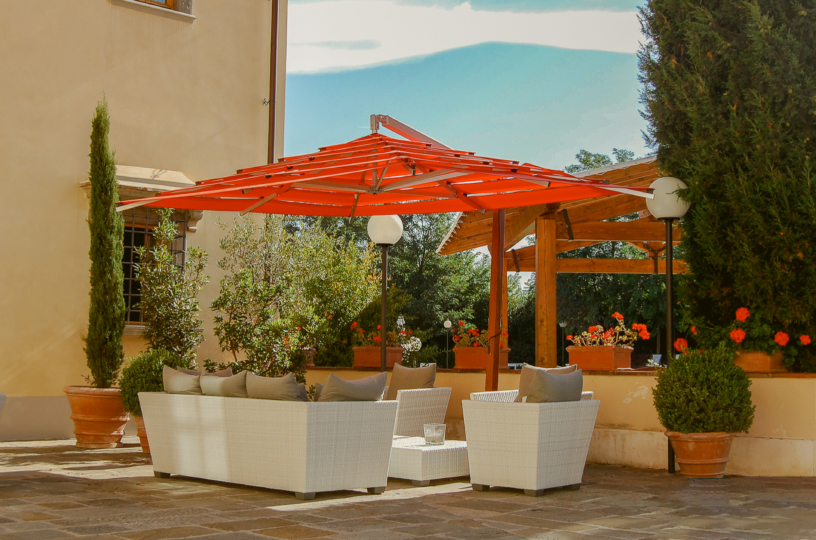 Outdoor patio with white wicker furniture under a red umbrella, surrounded by potted plants and trees.
