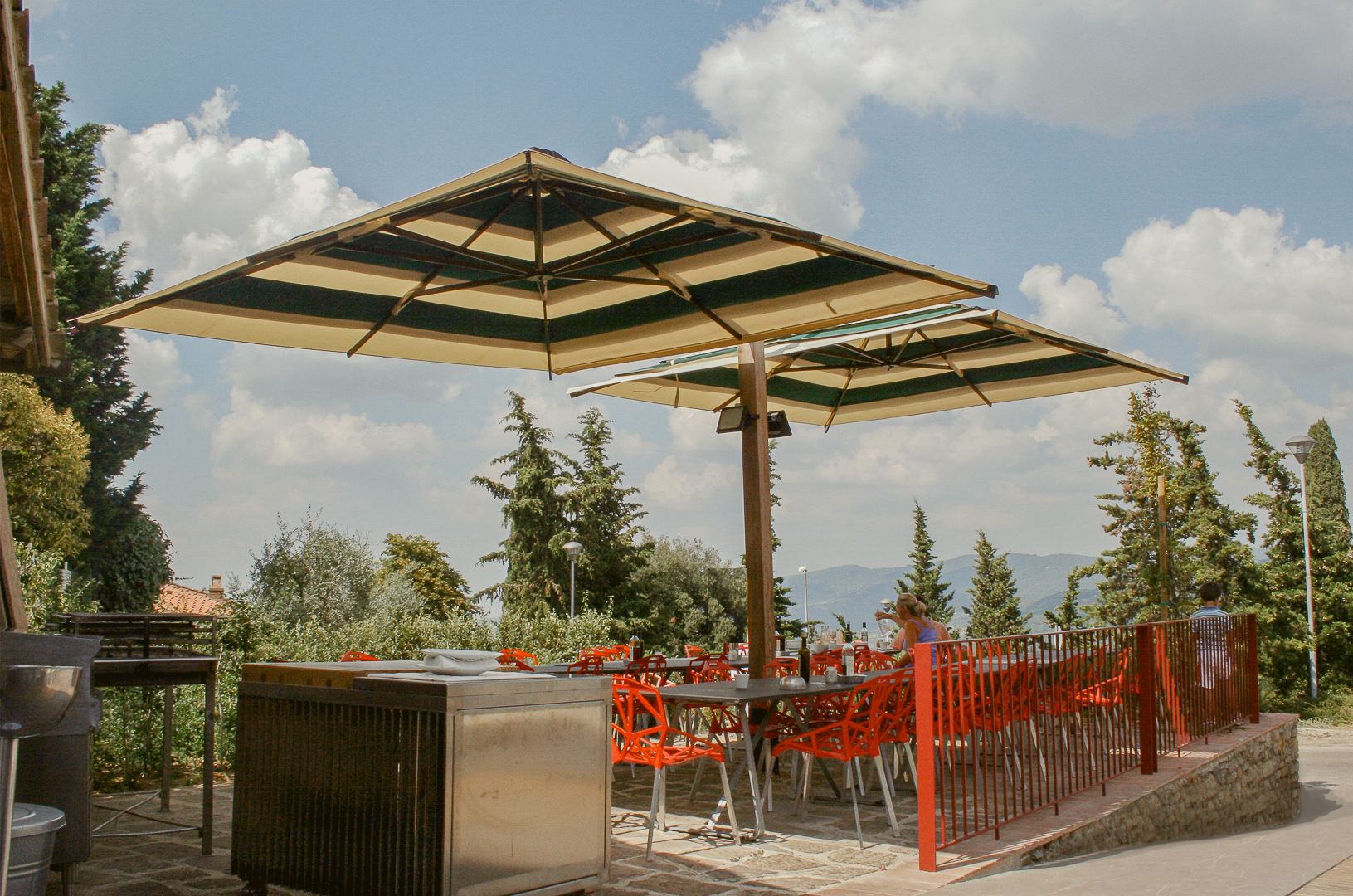Outdoor patio with red chairs, tables, and large striped umbrellas under a partly cloudy sky.