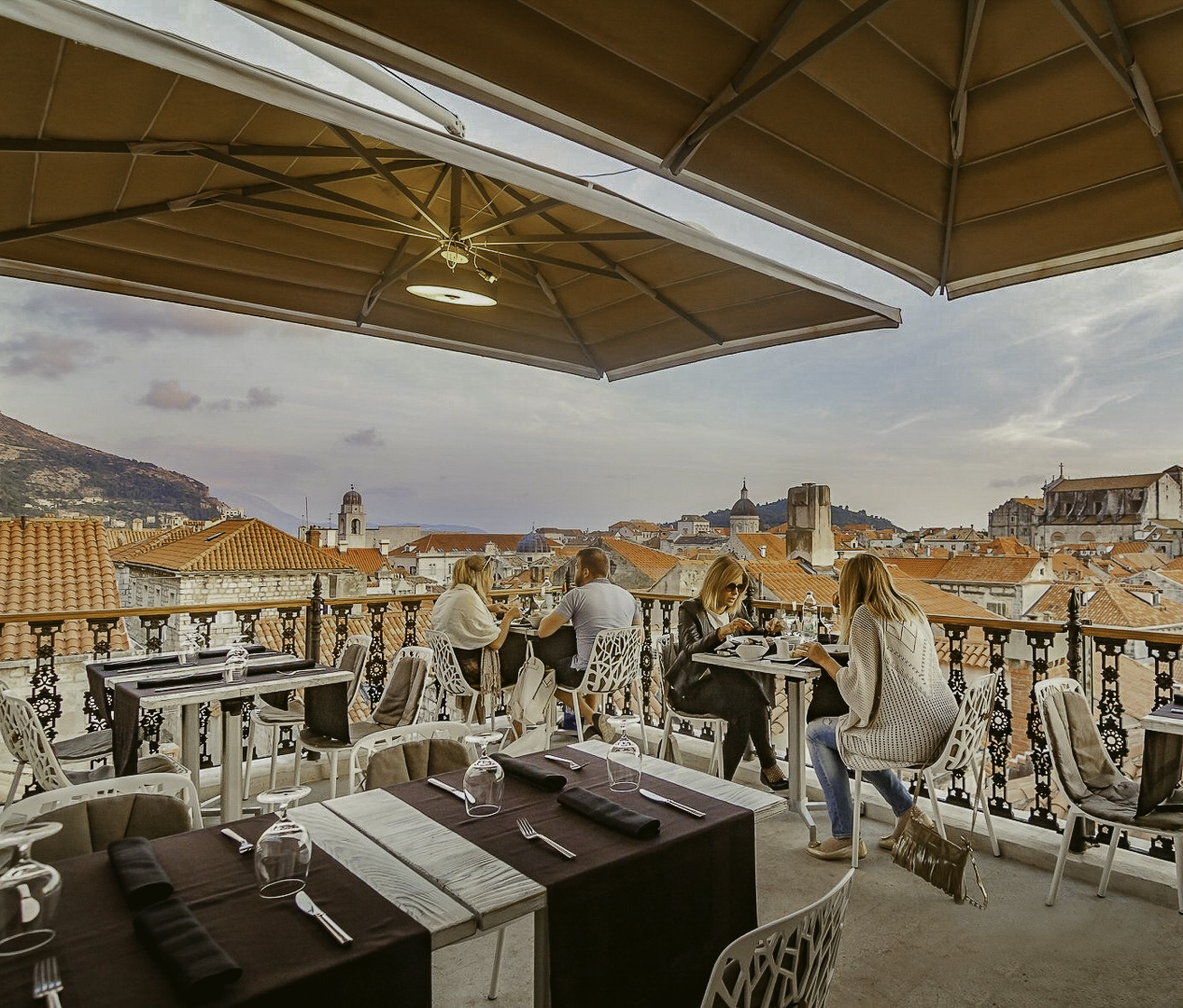 People dining on a rooftop terrace with views of orange rooftops and distant hills.