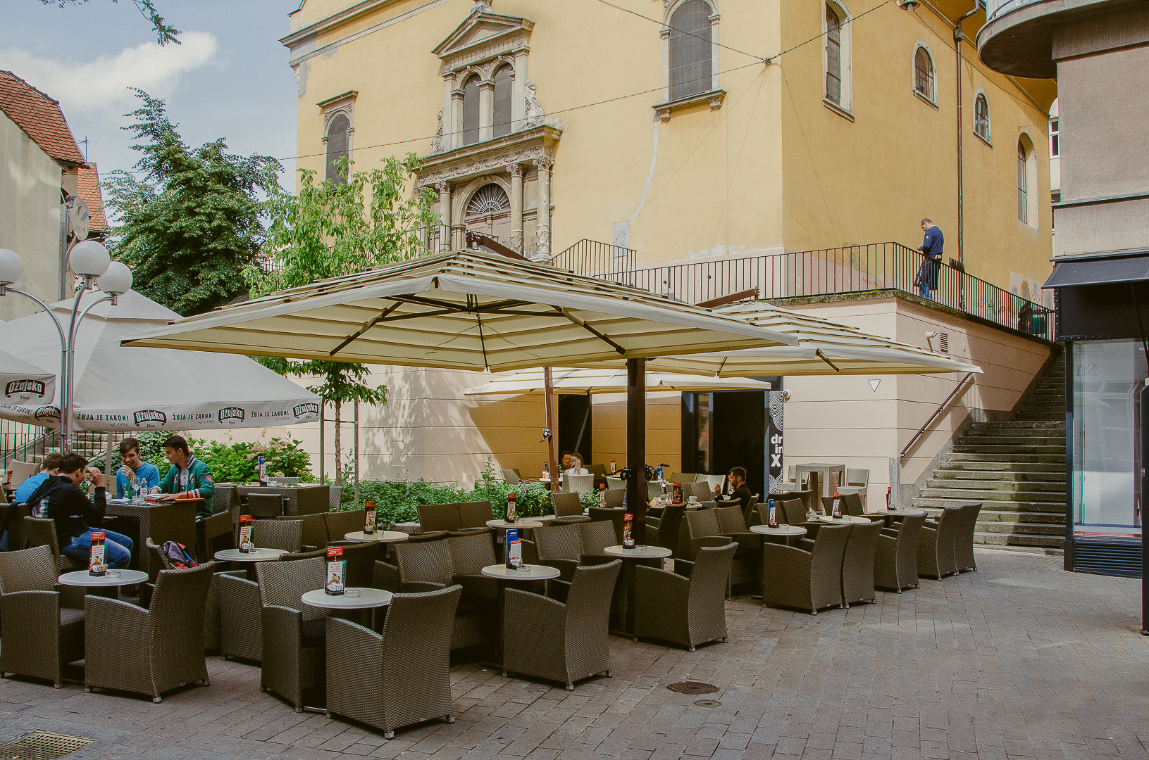 Outdoor cafe with empty wicker chairs and tables under large umbrellas, adjacent to a yellow building.