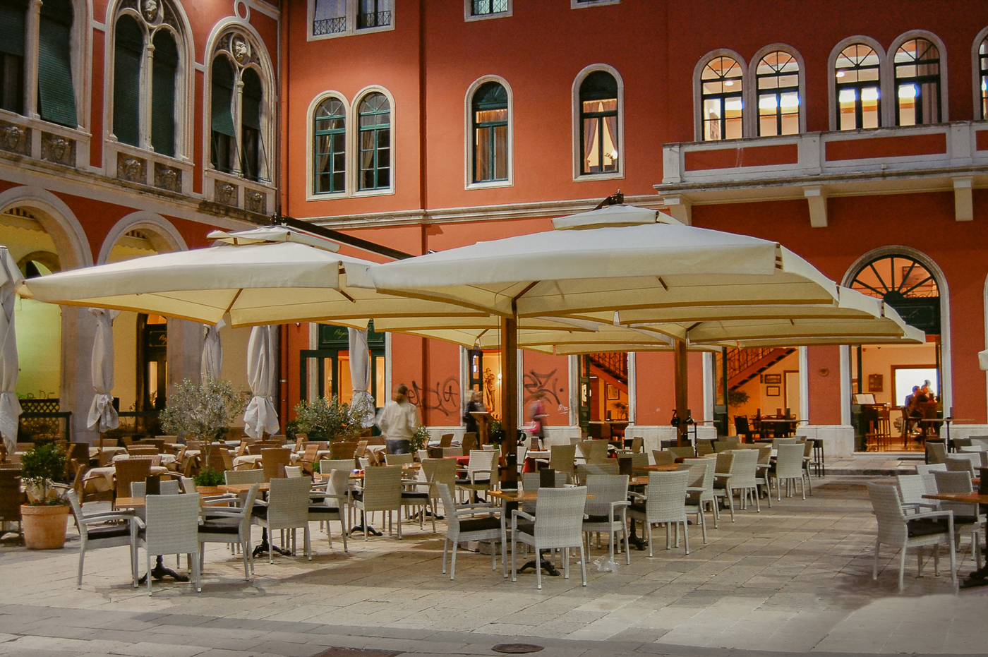 Outdoor café with white tables and chairs under large umbrellas in a courtyard.