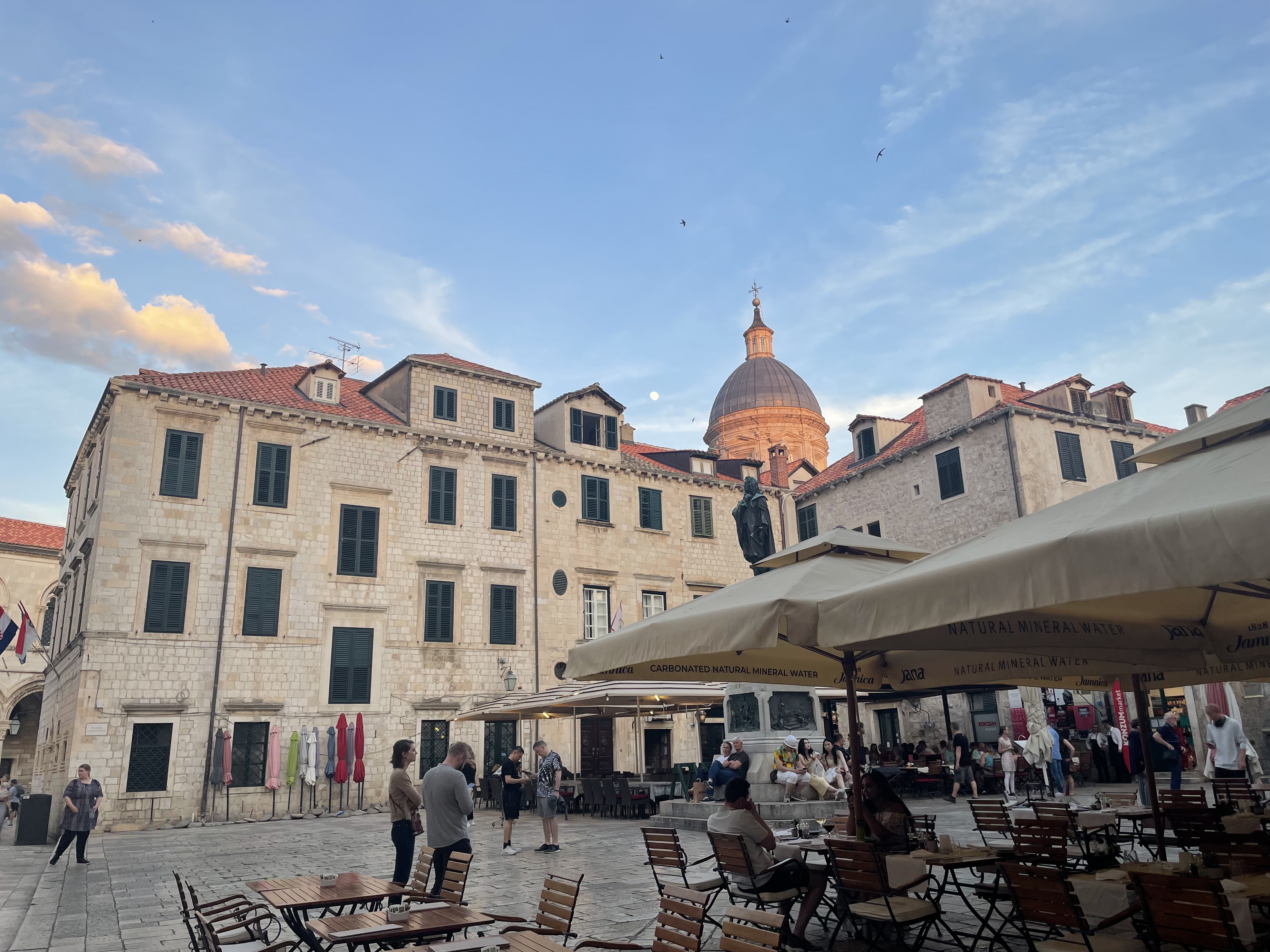 A plaza with outdoor seating, surrounded by old stone buildings and a church dome in the background.