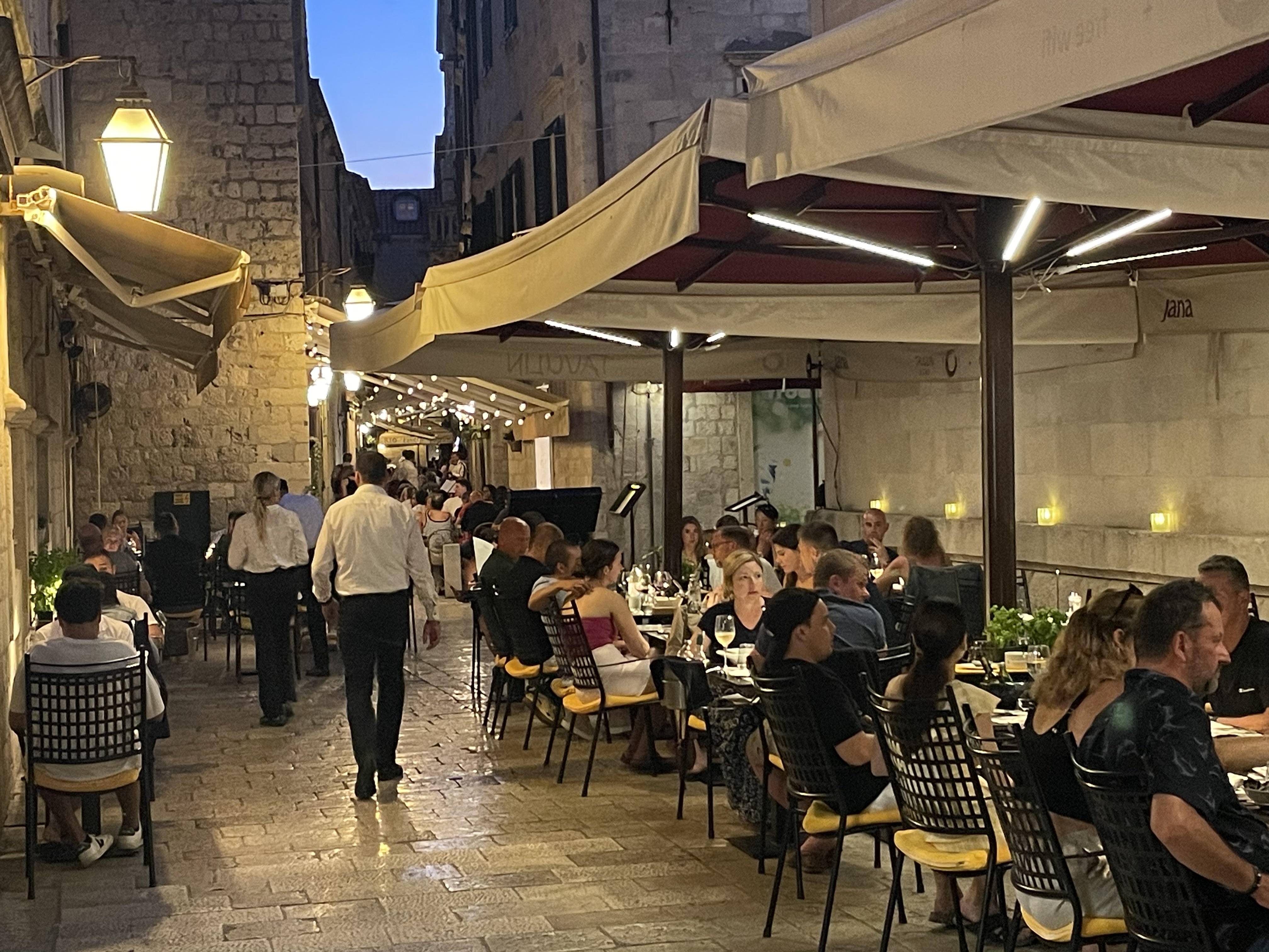 People dining at outdoor tables on a narrow, lit-up street in the evening.