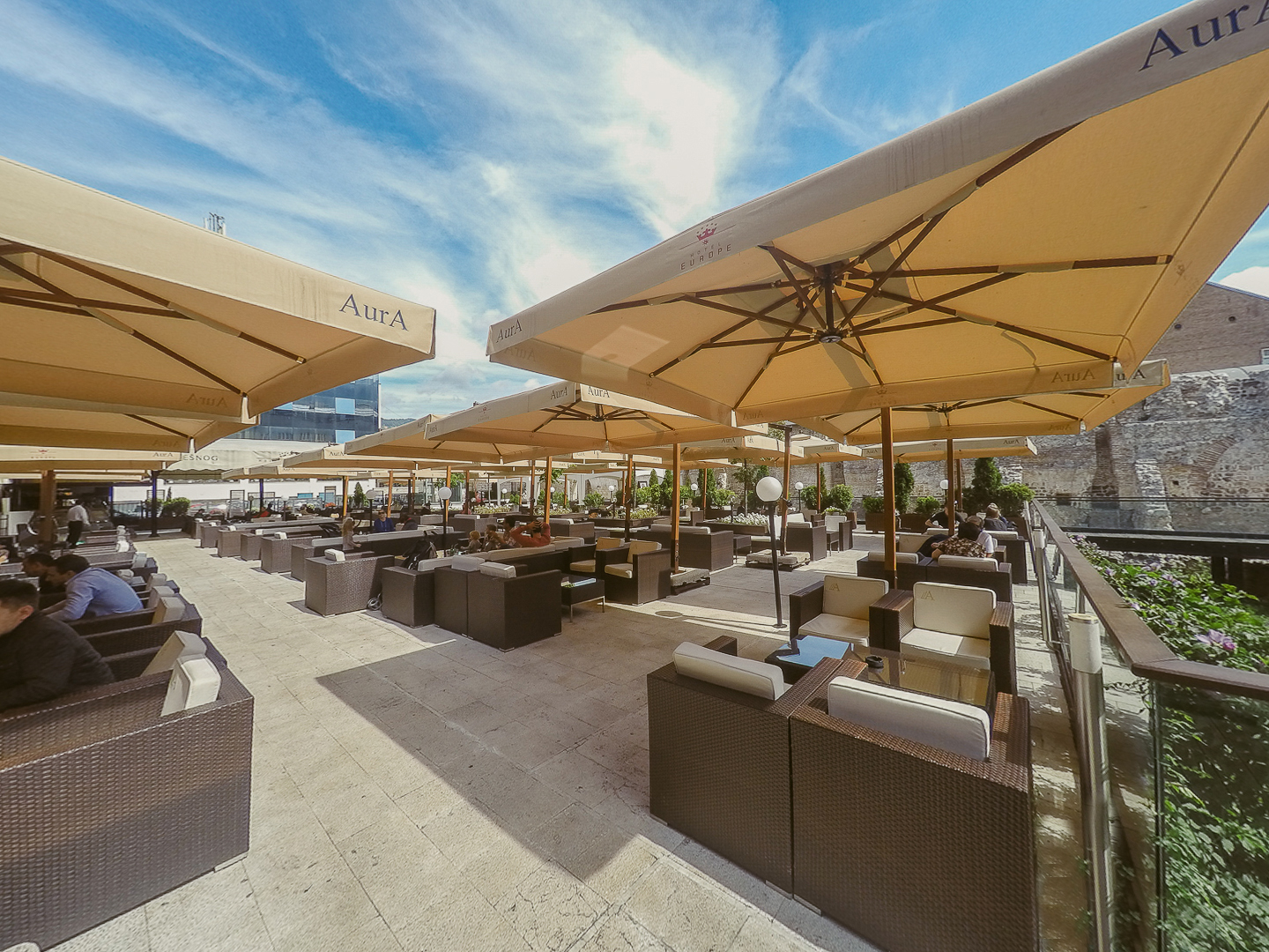Outdoor cafe with large umbrellas and wicker furniture, several people seated and dining.