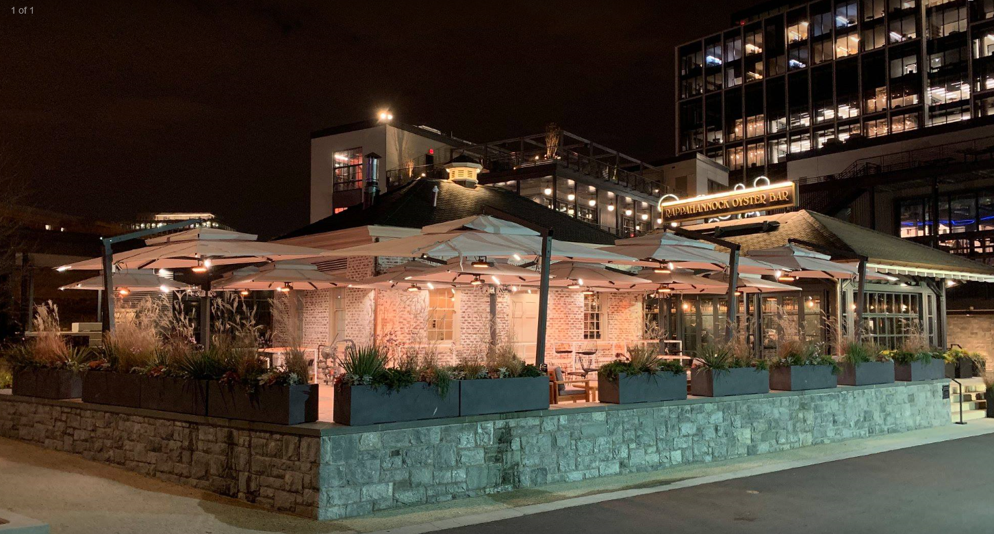 Outdoor restaurant seating area with large umbrellas, warm lighting, and heating systems at night.