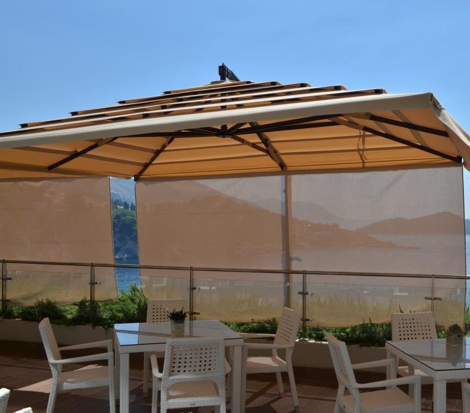 A patio with tables and chairs under a large beige canopy with side walls near a waterfront view.