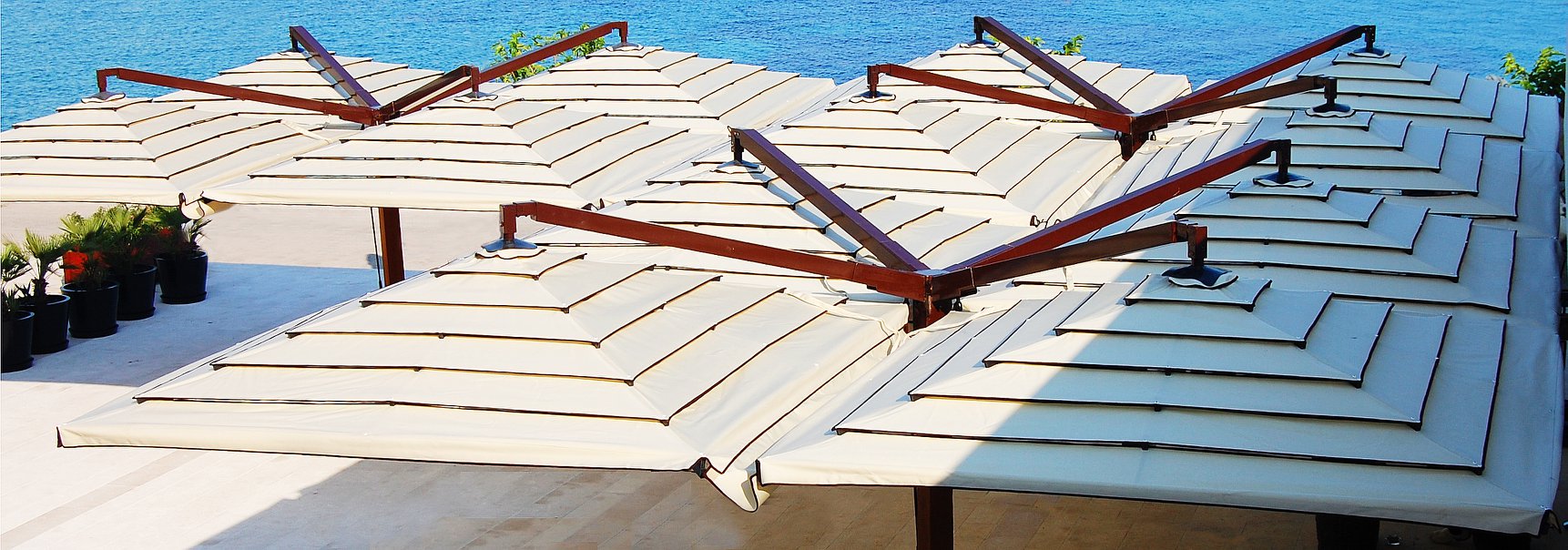 Large white umbrellas with wooden frames and rain gutter systems provide shade on a terrace near the ocean.