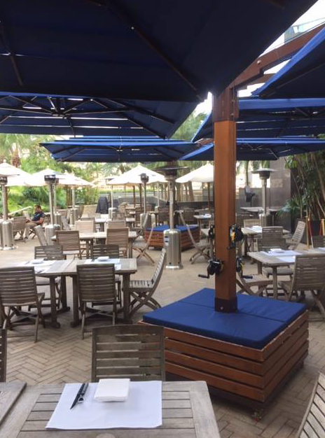 Outdoor restaurant seating with wooden tables and chairs under large blue umbrellas. No people are present.