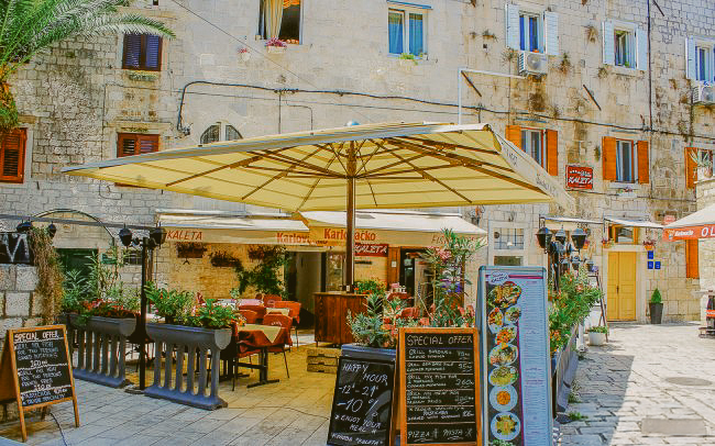 Outdoor restaurant with tables, chairs, and menus under large beige umbrellas against an old stone building.