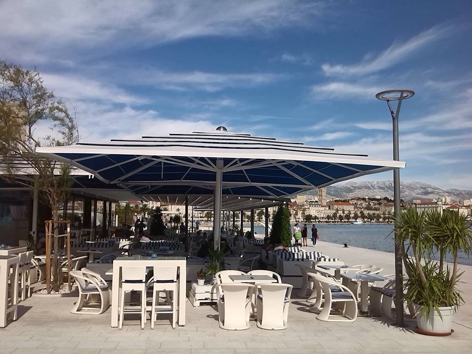 Outdoor café with white tables and chairs beneath large blue umbrellas on a sunny day.