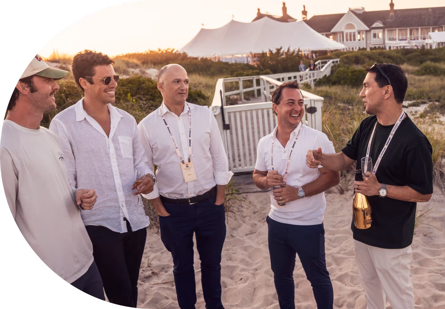 Five men standing on sand near a beach house, smiling and socializing at sunset, two holding drinks.
