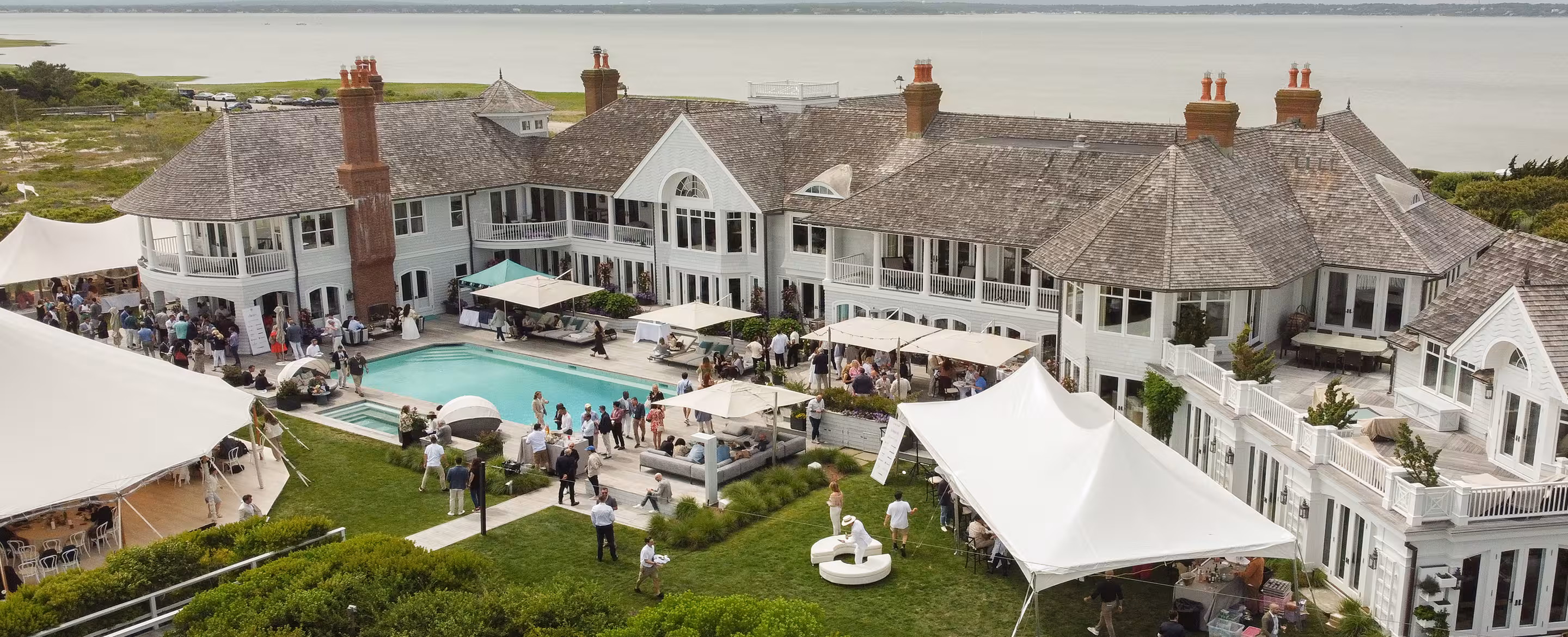 Large white mansion with decks, a pool, and tents on a lawn by a body of water during an outdoor event.