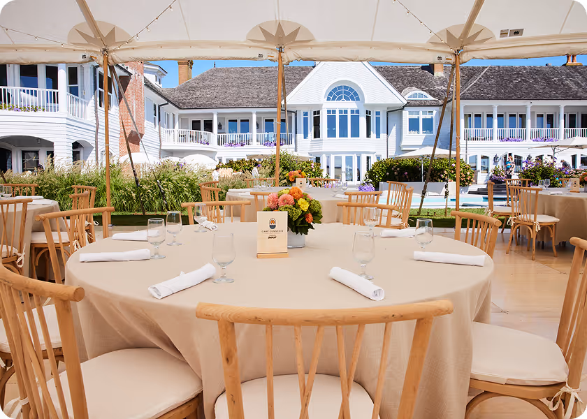 Outdoor dining tables with beige tablecloths and wooden chairs under a tent near a large white house.