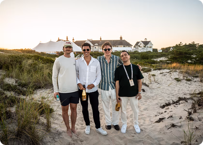 Four young men standing on sandy beach dunes holding drinks, with houses and tents in the background at sunset.
