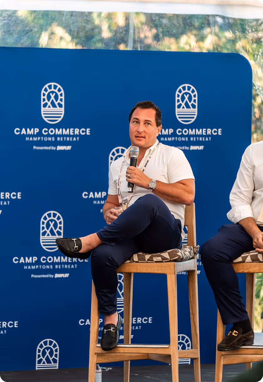 Man in white shirt speaking into microphone while seated on wooden stool at Camp Commerce Hamptons Retreat.