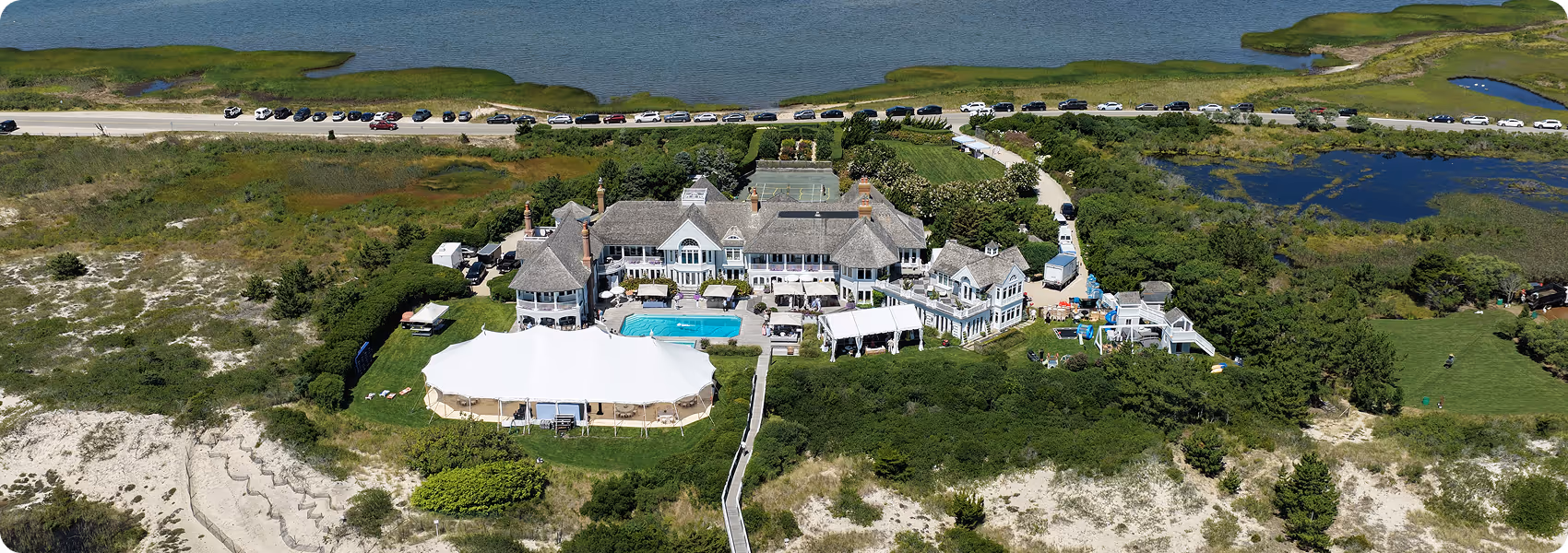 A large estate with a pool, white event tent, tennis court, and lush greenery near a body of water.