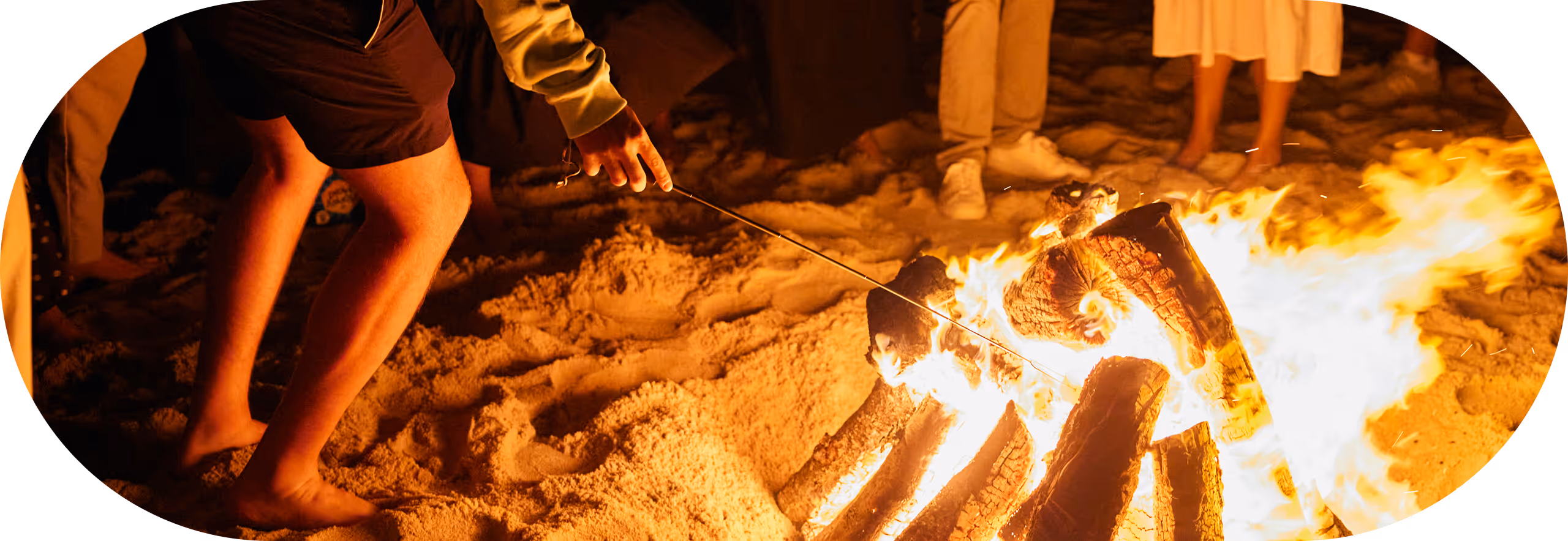 Group of people standing barefoot on sand around a bonfire with one person roasting marshmallows.