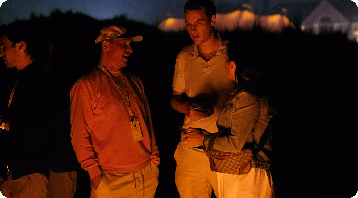 Four people talking and laughing around a campfire at night with dim warm light on their faces.