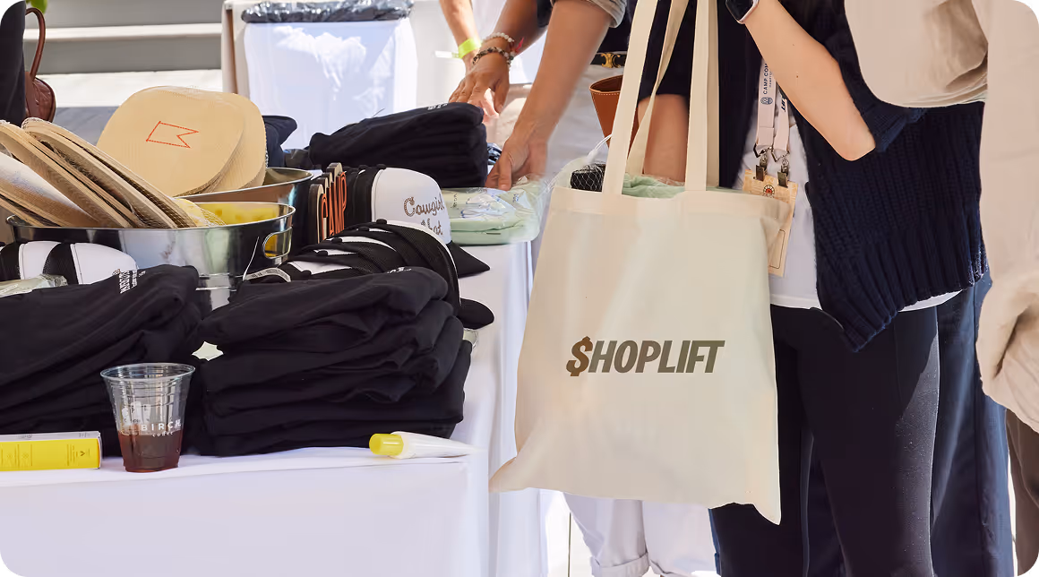People browsing and picking up merchandise at an outdoor event table with a tote bag labeled SHOPLIFT.