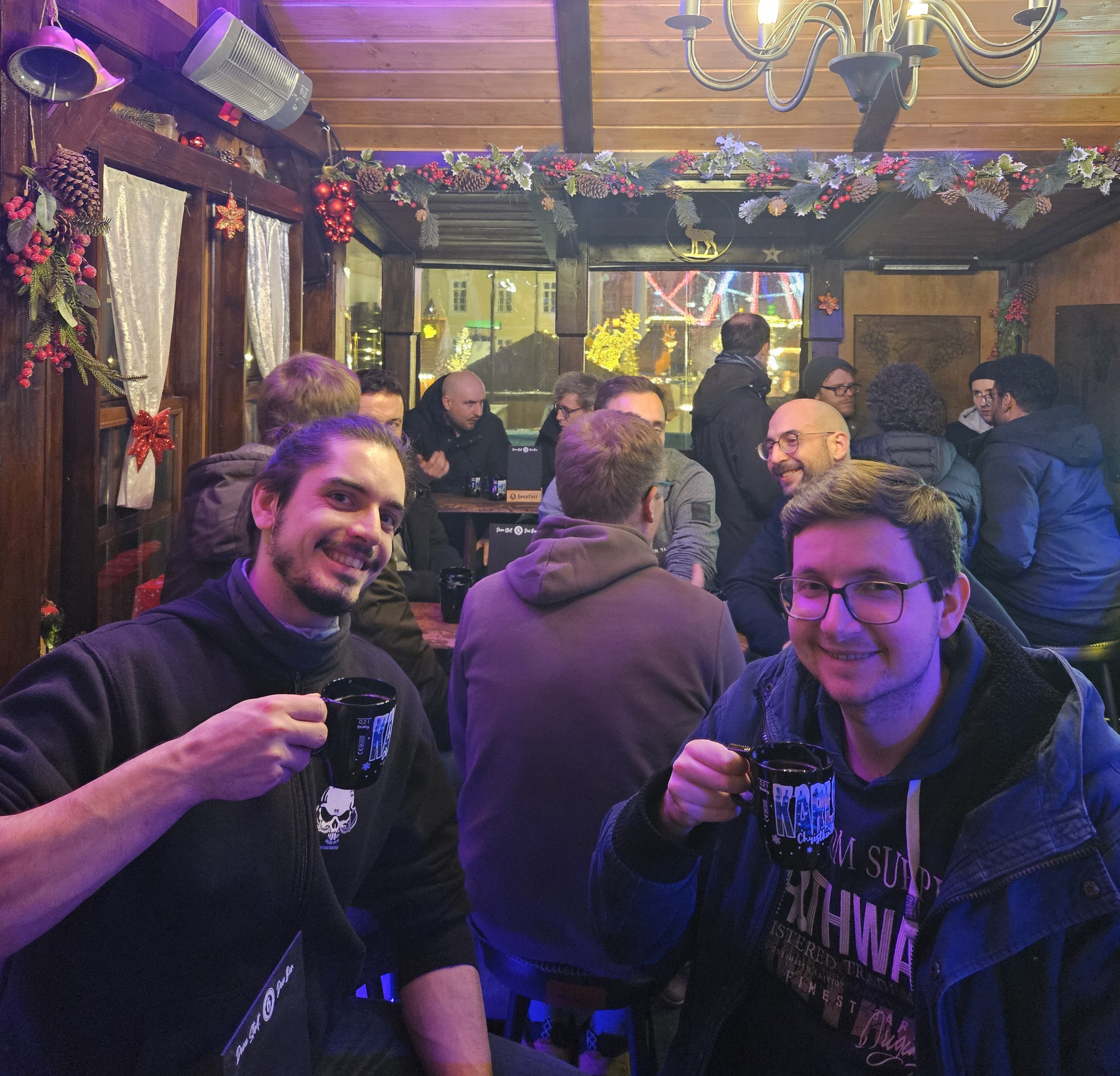 Zwei junge Männer in einer Hütte auf dem Weihnachtsmarkt prosten mit Glühweintassen freundlich in die Kamera