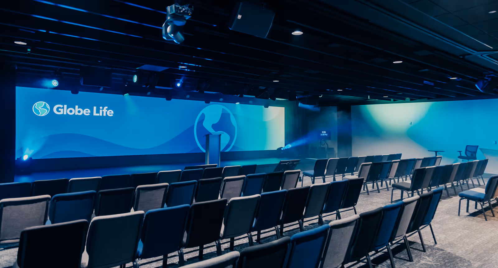 Contemporary auditorium stage with podium, large LED backdrop, and overhead speakers and stage lighting.