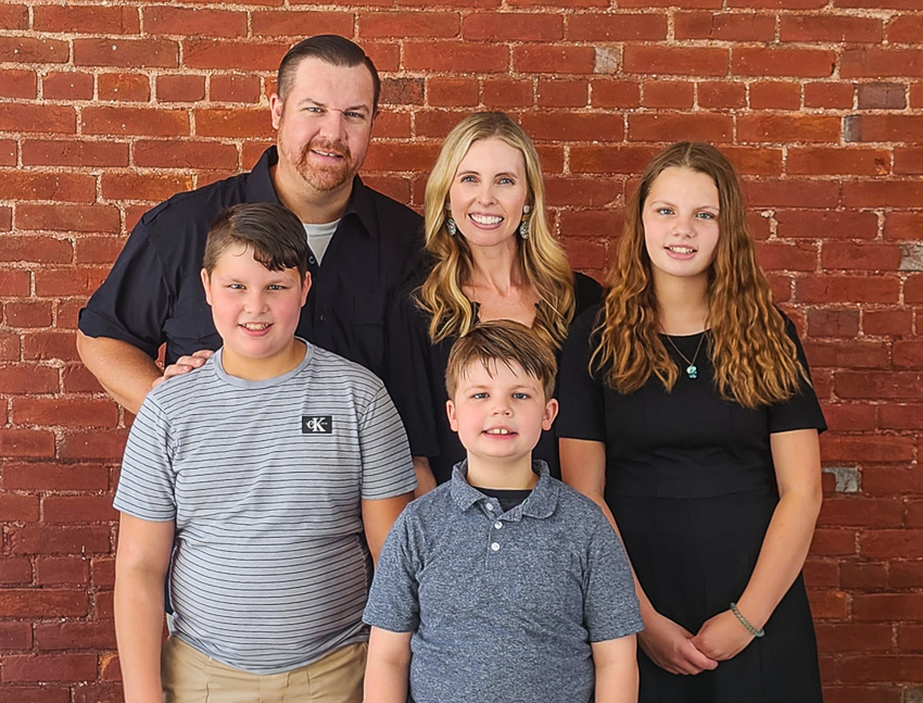 Group portrait of a smiling family of five standing in front of a red brick wall.
