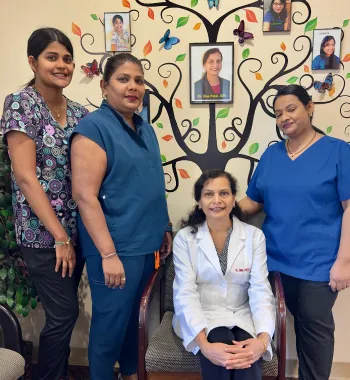 Four women medical staff posing in front of a wall decorated with a family tree and portraits, three standing and one seated in a white coat.